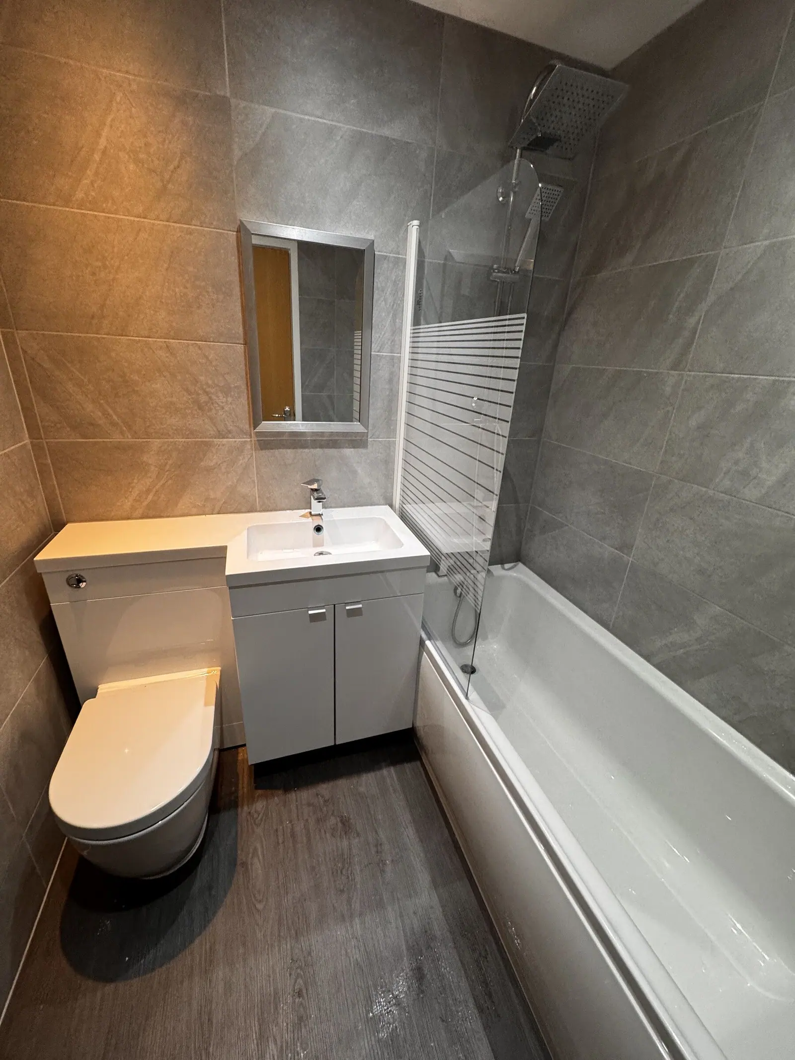 Compact bathroom with grey wall tiles, wood-effect floor, white vanity and WC unit and shower bath, fitted by Basingstoke Bathrooms